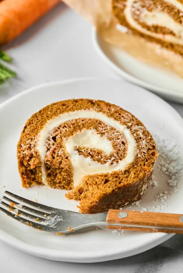 A slice of carrot cake roll with cream cheese filling on a white plate, beside a fork. A part of the roll is sliced off and a carrot is in the background.